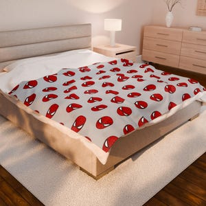 May include: A light gray blanket featuring a repeating pattern of red and white Spider-Man mask designs. The blanket is draped over a bed, showcasing the vibrant colors and detailed illustrations of the superhero's iconic mask.