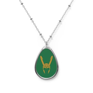 May include: Silver-toned necklace featuring a pendant with a green background and a gold-colored emblem. The pendant is oval-shaped and hangs from a delicate chain. The emblem is a stylized design, possibly a symbol or logo.
