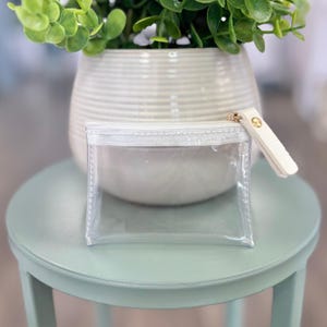 Pode incluir: Uma bolsa retangular transparente com zíper branco e uma pequena aba de couro branco sujo. A bolsa está em um banquinho verde claro, com um vaso branco e uma planta verde ao fundo.