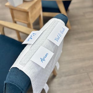 May include: A white fabric piece with a subtle dotted texture draped over a blue chair. Three white rectangular labels are placed on the fabric, featuring blue printed text that reads Robe, Veil & Sash, and Apron, identifying specific items.