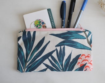 May include: A rectangular pencil case with a pink zipper and a botanical print of teal and dark blue leaves and coral flowers on a cream background. Pens and a card with text are visible inside the case.