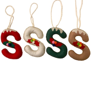 May include: Four felt Christmas decorations shaped like the letter 'S'. Each decoration is a different colour: red, white, green, and brown. Each decoration has a white icing design and colourful pom-poms.