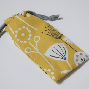 Peut inclure: Une pochette jaune à cordon de serrage avec un motif floral blanc, gris et noir. La pochette a une fermeture à cordon de serrage grise. Le tissu présente des fleurs et des têtes de graines stylisées.