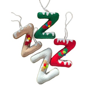 Könnte beinhalten: Vier Filz-Weihnachtsbaumschmuck in Form des Buchstabens Z. Jeder Schmuck ist in einer anderen Farbe: grün, beige, weiß und rot. Jeder Schmuck hat einen weißen Pompon und einen roten Pompon.