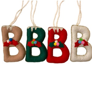 Könnte beinhalten: Vier Filz-Weihnachtsbaumschmuck in Form des Buchstabens B. Jeder Schmuck ist in einer anderen Farbe: beige, grün, rot und weiß. Jeder Schmuck hat einen Zweig mit roten Beeren und bunten Bommeln.