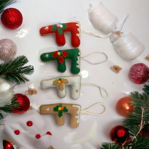 Könnte beinhalten: Vier Filz-Weihnachtsbaumschmuck in Form des Buchstabens 'T' in Rot, Grün, Weiß und Braun. Jeder Schmuck ist mit einem kleinen grünen Zweig und roten Beeren mit goldenen Glitzerakzenten verziert. Der Schmuck hat weiße Schnur-Schlaufen zum Aufhängen.