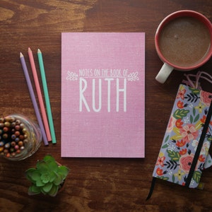 May include: A pink notebook with the text Notes on the Book of Ruth printed in white, accompanied by three colored pencils, a jar of colored pencils, a small succulent plant, a floral patterned notebook with an elastic band, and a mug of coffee.