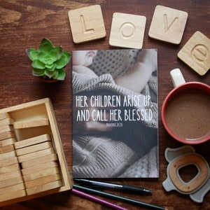 Puede incluir: Un libro con el texto "Sus hijos se levantan y la llaman bendita" y una referencia a un versículo bíblico. El libro está abierto en una página con una foto de un bebé durmiendo en una manta. El libro está rodeado de bloques de madera que deletrean la palabra "love".