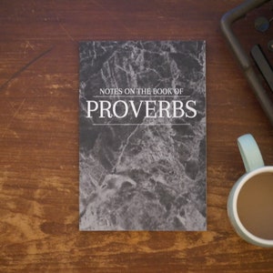 Puede incluir: Un libro con cubierta de mármol en blanco y negro con el título "Notes on the Book of Proverbs". El libro está sobre una mesa de madera con una máquina de escribir vintage y una taza de café.