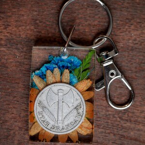 Può includere: Un portachiavi argentato con un ciondolo in resina trasparente che presenta una disposizione di fiori pressati e una moneta d'argento con il testo "ETAT FRANCAIS" e un design di ascia.