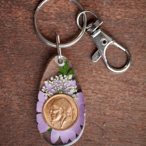 Könnte beinhalten: Ein silberner Schlüsselanhänger mit einem klaren Harz-Anhänger, der eine gepresste Blume und eine goldene Münze zeigt. Die Münze zeigt das Profil einer Person.
