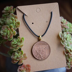 Może przedstawiać: Naszyjnik z miedzianym wisiorem w kształcie dwupensowej monety z wizerunkiem lwa. Moneta jest na czarnym sznurku i jest prezentowana na brązowej kartce.