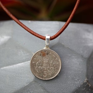 Puede incluir: Un collar de cordón de cuero marrón con un colgante de plata que presenta una moneda británica de cinco peniques. La moneda tiene un escudo real y la inscripción "FIVE PENCE".