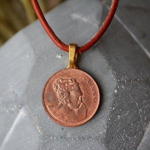 Puede incluir: Un collar con un colgante de color cobre que muestra la imagen de perfil de un rey con una corona. El colgante está inscrito con la palabra "CADNABO". El collar está hecho de cordón de cuero marrón.