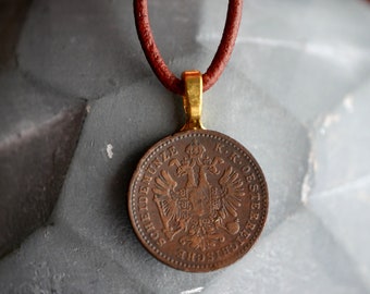 Könnte beinhalten: Eine braune Lederkordelkette mit einem goldfarbenen Anhänger, der eine Kupfermünze mit der Aufschrift "SOHLIDE MUNZE K.K.OESTERREICH" und ein Bild eines Adlers mit einem Schild zeigt.