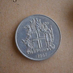 May include: A silver coin featuring a detailed embossed design. The coin displays a coat of arms with a shield, figures, and the year 1967. The coin is round and has a slightly worn appearance, suggesting it is vintage or antique.