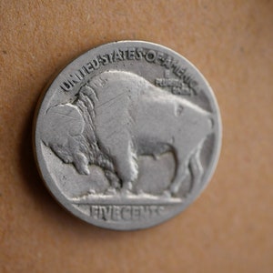 May include: A silver US five-cent coin featuring a profile of a buffalo with the words "UNITED STATES OF AMERICA" and "FIVE CENTS" inscribed around the edge.