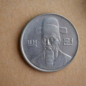 May include: A silver Korean Won coin featuring a portrait of King Sejong the Great, the inventor of the Korean alphabet, Hangeul. The coin has the Korean word "Won" written on it.