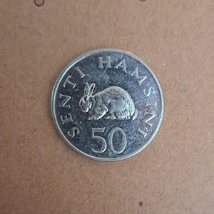 以下が含まれることがあります： 詳細なウサギの描写が施された銀貨。コインの縁には「HAMSIM」と「SENTI」の文字が刻まれ、数字の「50」が大きく表示されています。茶色の背景に置かれたコインです。