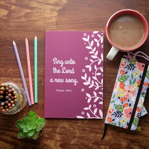 Könnte beinhalten: Ein violettes Notizbuch mit dem Text "Sing unto the Lord a new song. Psalm 149:1" auf dem Einband. Das Notizbuch ist umgeben von bunten Bleistiften, einer Tasse Kaffee und einem Notizbuch mit Blumenmuster.