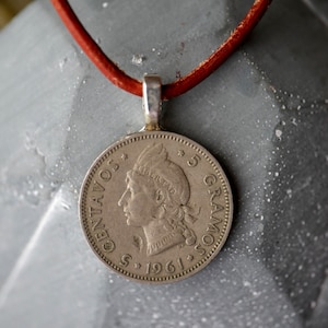 Puede incluir: Un collar con un colgante de moneda de plata y un cordón de cuero rojo. La moneda presenta el perfil de una mujer con el texto "5 CENTAVOS" y "5 GRAMOS" alrededor del borde. La fecha "1961" también es visible en la moneda.