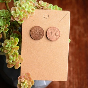 May include: A pair of copper-colored earrings featuring a vintage coin design with a royal crest. The earrings are displayed on a brown card with a hole for hanging.