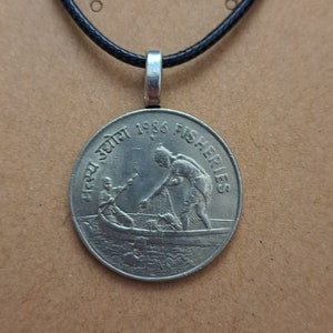 May include: A silver coin pendant with a black cord. The coin features an image of two people fishing from a boat, with the text "1986 FISHERIES" and other text in a foreign language.