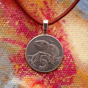 May include: A silver pendant necklace featuring a five dollar New Zealand coin with an image of a lizard on it. The coin is on a brown leather cord.