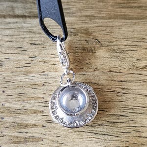 May include: A silver-toned metal charm shaped like a small teacup and saucer, featuring a detailed embossed pattern along the rim of the saucer. The charm is attached to a small silver lobster clasp, which is connected to a black zipper pull.