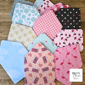 May include: A collection of assorted paper envelopes featuring various patterns including pink flamingos, cassette tapes, hearts, stars, clouds, polka dots, and floral designs. A white square label in the corner displays the text Pretty Charms.