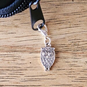 May include: A silver-toned metal owl charm attached to a black zip pull by a small lobster clasp. The owl charm features engraved details representing eyes, feathers, and wings, hanging vertically from the zip mechanism of a dark item.