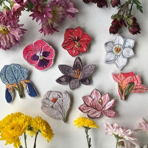 Könnte beinhalten: Eine Sammlung von gestickten Blumen-Patches in verschiedenen Farben, darunter Rot, Pink, Weiß und Blau. Die Patches sind alle in unterschiedlichen Formen und Größen und in einem zufälligen Muster auf einem weißen Hintergrund angeordnet.