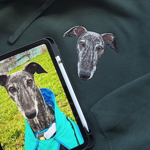 Puede incluir: Sudadera con capucha verde oscuro con una cabeza de galgo bordada en el pecho. Una tableta muestra una foto de un galgo con un abrigo azul. Un lápiz óptico blanco descansa sobre la tableta. La sudadera tiene una capucha con cordón.