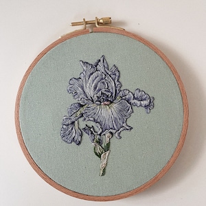 Può includere: Un fiore di iris ricamato in viola e verde, esposto in un cerchio di tessuto verde chiaro.