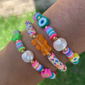 May include: A selection of colourful beaded bracelets. Each bracelet features a variety of beads, including round pearls, striped cylinders, and flower-shaped beads. Some bracelets include a gummy bear charm and an evil eye charm. The bracelets are strung on a stretchy cord.
