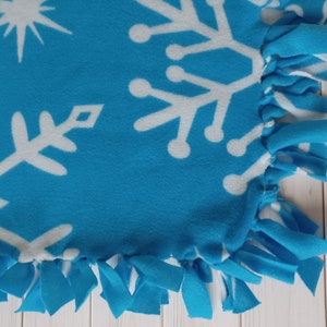 Könnte beinhalten: Eine blaue Fleece-Decke mit weißen Schneeflocken-Designs. Die Decke hat einen Fransenrand.