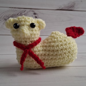 May include: A cream-coloured, crocheted animal figure with black button eyes and a red scarf. A small red crocheted detail, possibly a tail or wing, is attached to the back of the figure.