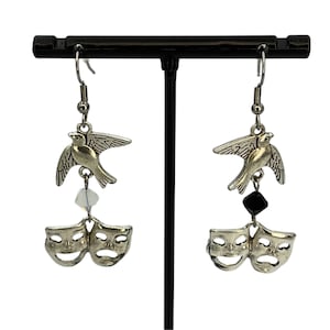 Op de afbeelding: Zilverkleurige oorhangers met een vogelbedel boven komedie- en tragediemaskers. Eén oorbel heeft een heldere kraal, de andere een zwarte kraal. De oorbellen hangen aan een zilveren haak.