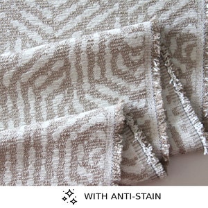 Op de afbeelding: Close-up van een witte stof met een bruin geometrisch patroon. De stof heeft een gefranst randje en de tekst "WITH ANTI-STAIN" is zichtbaar onderaan de afbeelding.