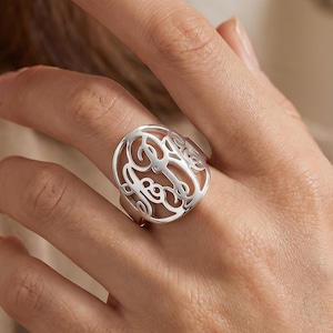 Peut inclure: Une bague argentée avec un motif ajouré représentant des initiales entrelacées. La bague est ronde et portée au doigt. Le design est complexe et détaillé, avec une finition polie.
