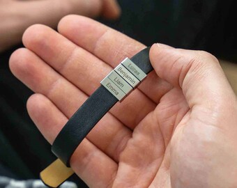 May include: A black leather bracelet with silver name bars. The bracelet is held in a person's hand. The silver bars are engraved with the names: Lunas, Benjamin, Liam, and Emma.