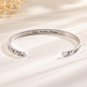 May include: A silver-colored cuff bracelet with the engraved words "Give. Thanks. Always." The open-ended bracelet features a floral design on both ends. The bracelet is displayed on a light-colored surface.