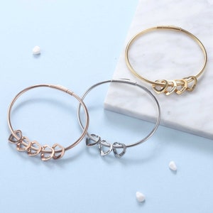 May include: Three silver bangle bracelets with heart-shaped charms. Each charm has a different name engraved on it: Emma, Lucas, Olivia, Ava, and Liam.