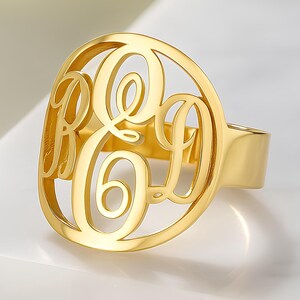 Könnte beinhalten: Ein goldfarbener Monogrammring mit den Buchstaben R, E und D in einem stilvollen, durchbrochenen Design. Der Ring hat einen glatten, polierten Ring und eine große, ovale Oberfläche mit den Initialen. Der Ring wird auf einer weißen Oberfläche präsentiert.