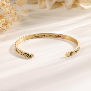 May include: A gold-colored cuff bracelet with engraved names: Emma, Noah, Liam, and Charlotte. The open-ended bracelet features a leaf design on both ends. The jewelry is displayed on a white surface.