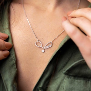 May include: Silver necklace with a stethoscope pendant and the name "Mila" inscribed on it. The necklace is worn by a person with a green shirt.