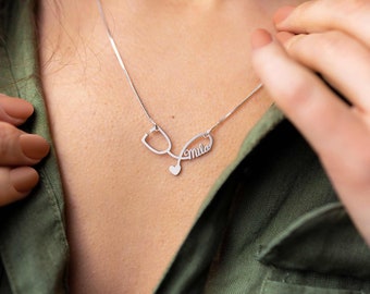 May include: Silver necklace with a stethoscope pendant and the name "Mila" inscribed on it. The necklace is worn by a person with a green shirt.