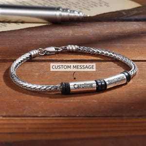 Op de afbeelding: Een zilveren gevlochten armband met een sluiting. De armband heeft twee cilindrische kralen met de namen "Caroline" en "Benjamin" gegraveerd. De tekst "CUSTOM MESSAGE" is ook zichtbaar.