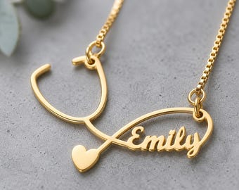 May include: A gold-toned necklace featuring a pendant shaped like a stethoscope with a small heart at the base. The name Emily is integrated into the design in a cursive script font. The pendant is attached to a delicate gold-toned box chain.