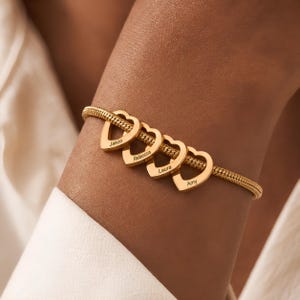Pode incluir: Uma pulseira de corrente de cobra em tom dourado com quatro berloques pendentes em forma de coração. Cada coração está gravado com um nome: Jason, Rebecca, Laura e Amy. A pulseira tem um acabamento polido e um design de elos trançados.